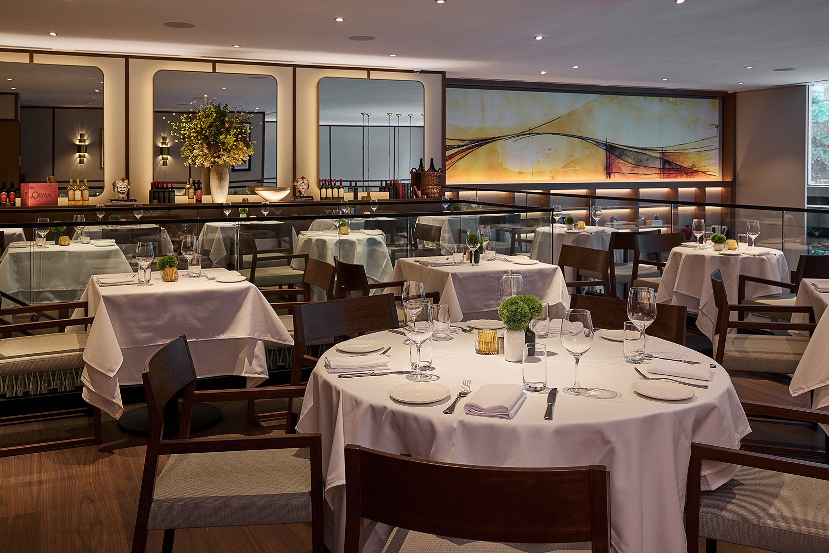 The interior of a high-end restaurant showing various seating arrangements, crisp white linens, and a large colorful mural behind a glass-railed mezzanine.