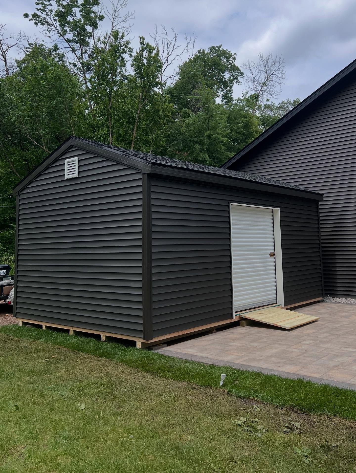 A newly installed, finished storage shed with dark gray vinyl siding and black trim sits on a wooden foundation in a backyard. It features a white roll-up door, a vent, and a small wooden ramp onto a paver patio.
