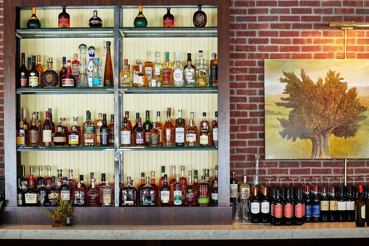 An organized display of premium spirits on glass shelves set against a cream background. To the right, a textured painting of an olive tree hangs on a red brick wall.