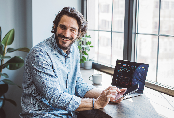 A man smiling while reviewing financial data on both his laptop and tablet.