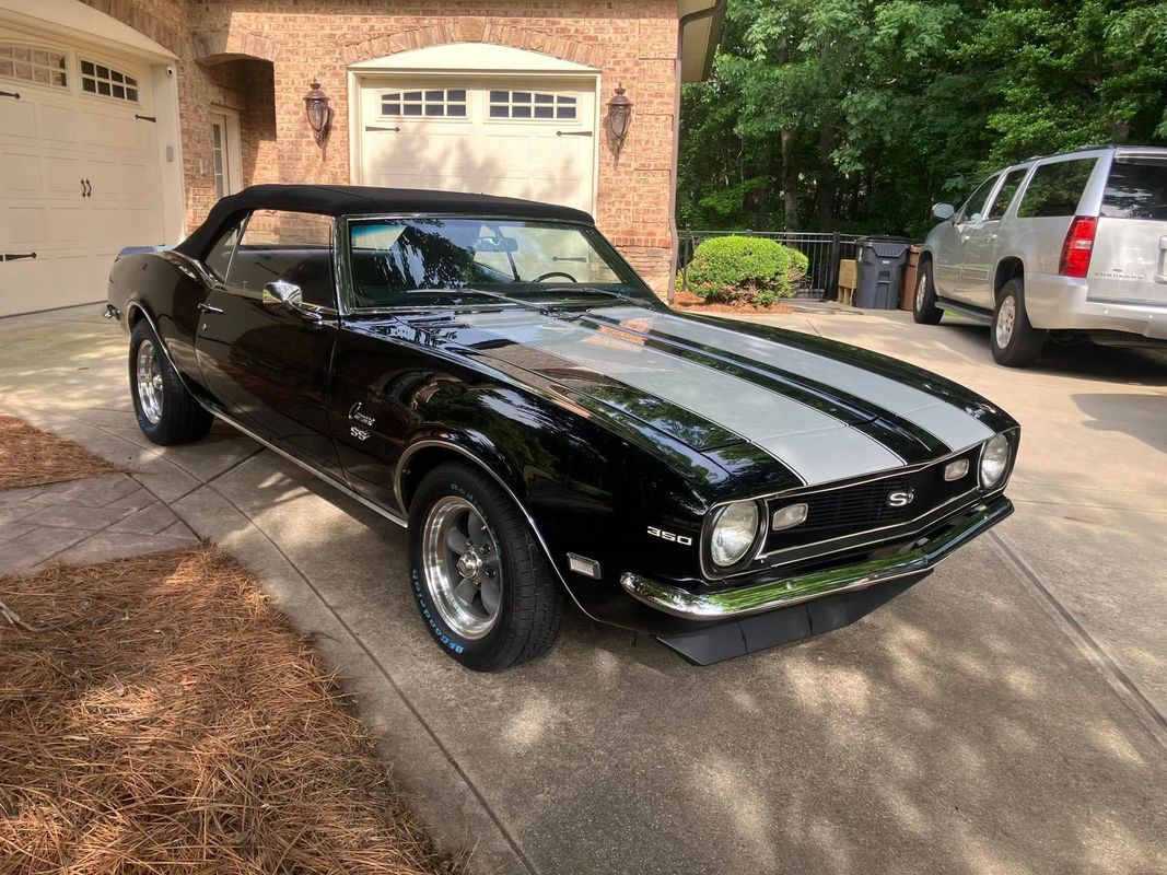 A classic Camaro convertible displays a deep black finish with contrasting silver stripes. The restoration highlights expert paintwork, body refinishing, and attention to automotive detailing quality.