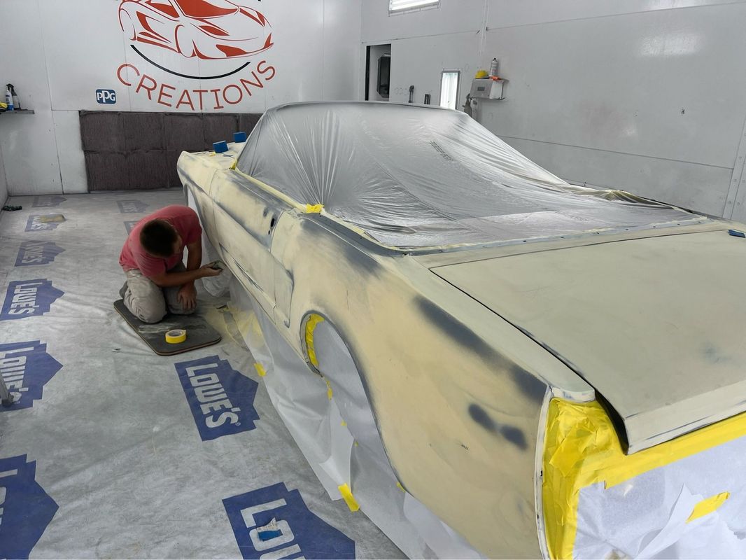 A worker carefully sands the body of a classic convertible during preparation for painting. The vehicle is masked and protected inside a clean spray booth designed for automotive refinishing.