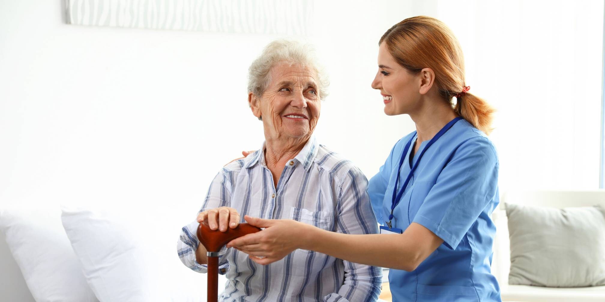 A caregiver in a blue uniform smiles while placing a hand on the shoulder of an elderly woman. The elderly woman, who has white hair and is wearing a striped shirt, is holding a wooden cane and looking at the caregiver. They appear to be in a well-lit room with white walls and furniture.