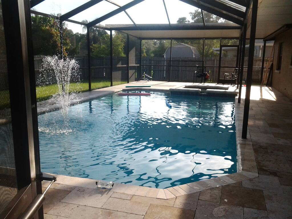 Screen-enclosed residential swimming pool with clear blue water, decorative fountain jets, and stone paver decking.
