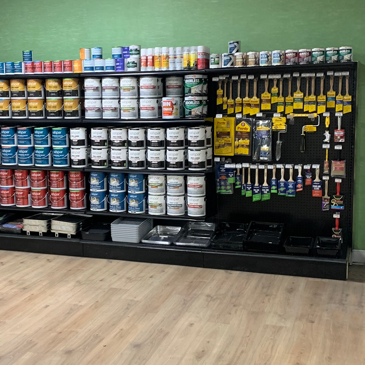 A neatly organized retail display shows rows of Valspar paint cans on black shelving beside a black pegboard filled with Purdy paint brushes, rollers, scrapers, and other painting tools. Paint cans are arranged by color, including blue, red, white, green, and neutral labels. Several paint trays are stacked along the bottom shelf on a light wood-look floor.