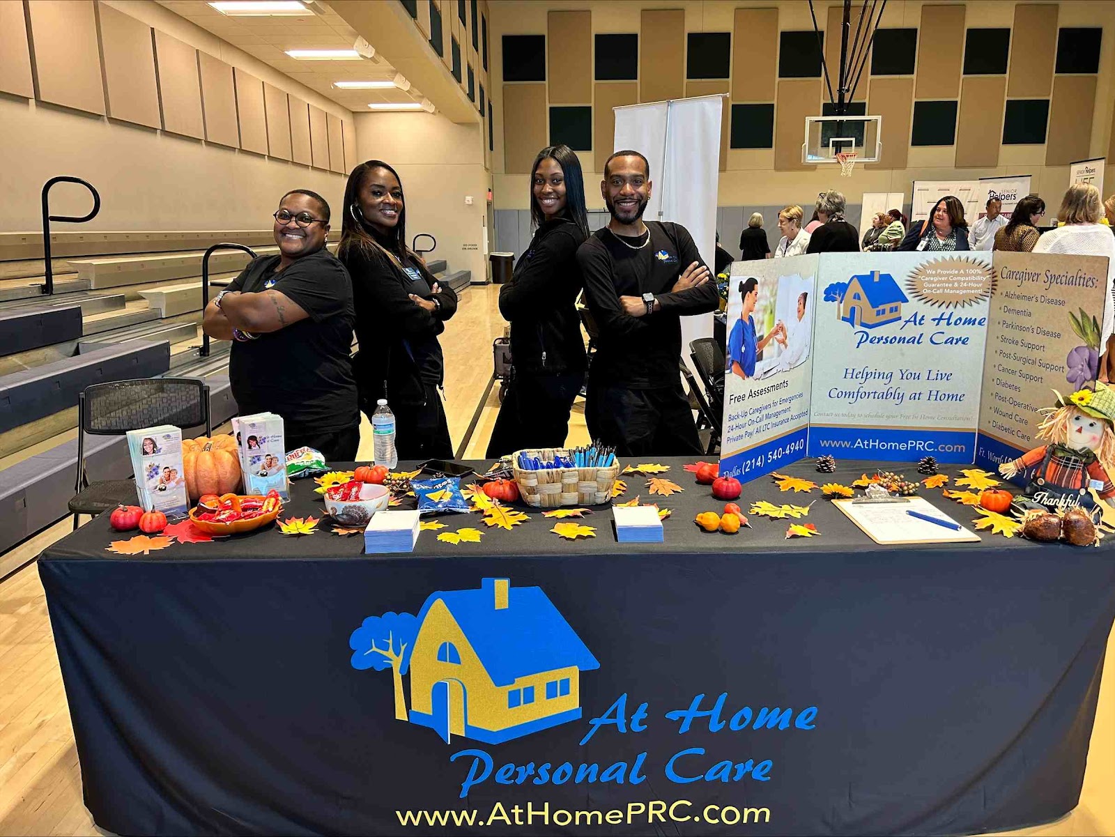 A group of four people stand behind a table at what appears to be a community event or health fair. The table is covered with a black tablecloth featuring the 'At Home Personal Care' logo. The table is decorated with fall-themed items like pumpkins, gourds, and artificial leaves. Several brochures and a sign listing caregiver specialties are displayed on the table. The people behind the table are smiling and appear to be representing the company. In the background, there are bleachers and a basketball hoop, suggesting the event is taking place in a gymnasium. Other attendees are visible in the background.