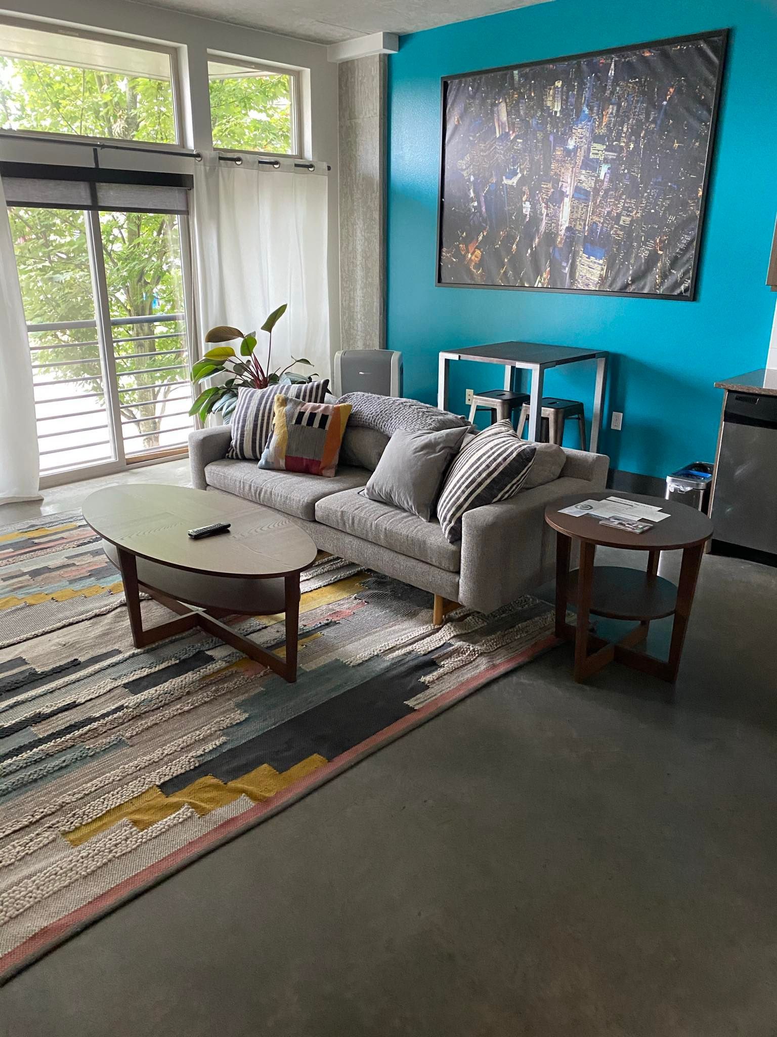 A modern living room featuring a gray sofa with colorful pillows, wooden coffee table, patterned rug, teal accent wall, large cityscape artwork, and bright windows creating a clean, stylish, inviting interior space.