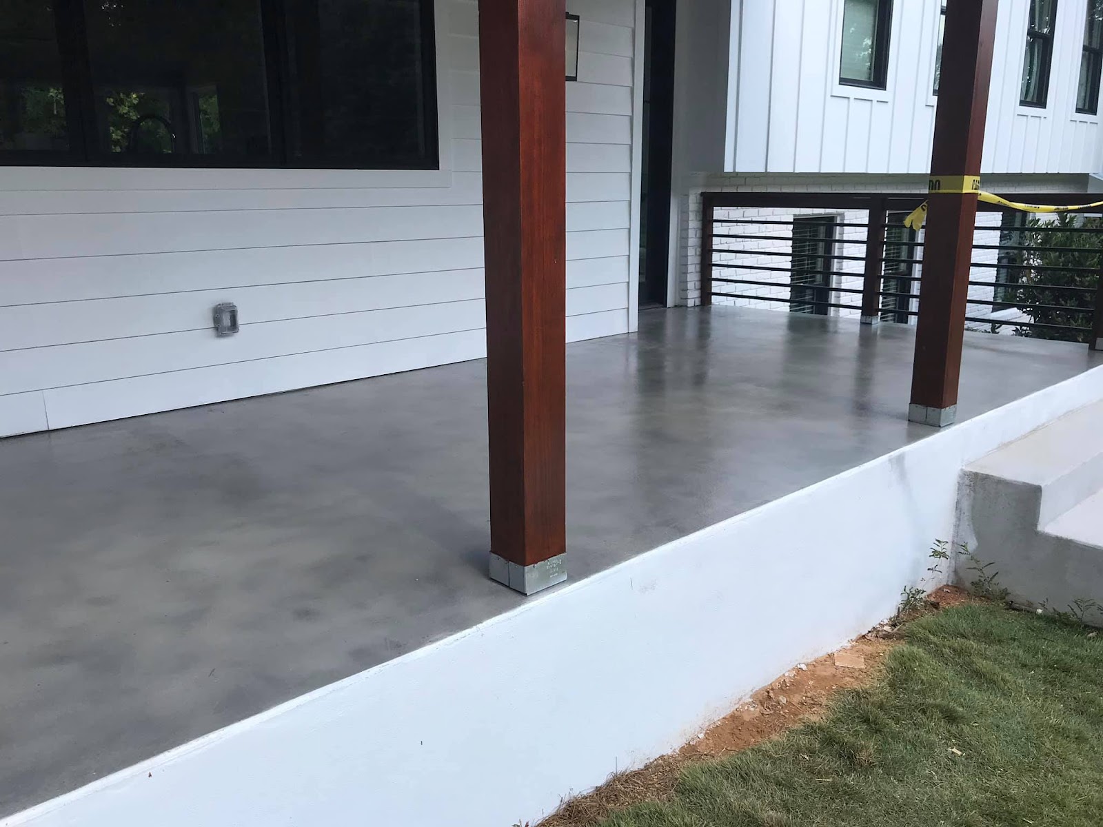 A modern porch with a polished concrete floor, white horizontal siding, and dark-framed windows. Two large wooden posts with silver bases support the roof. A black metal railing is visible in the background, and yellow caution tape is strung across one of the posts.
