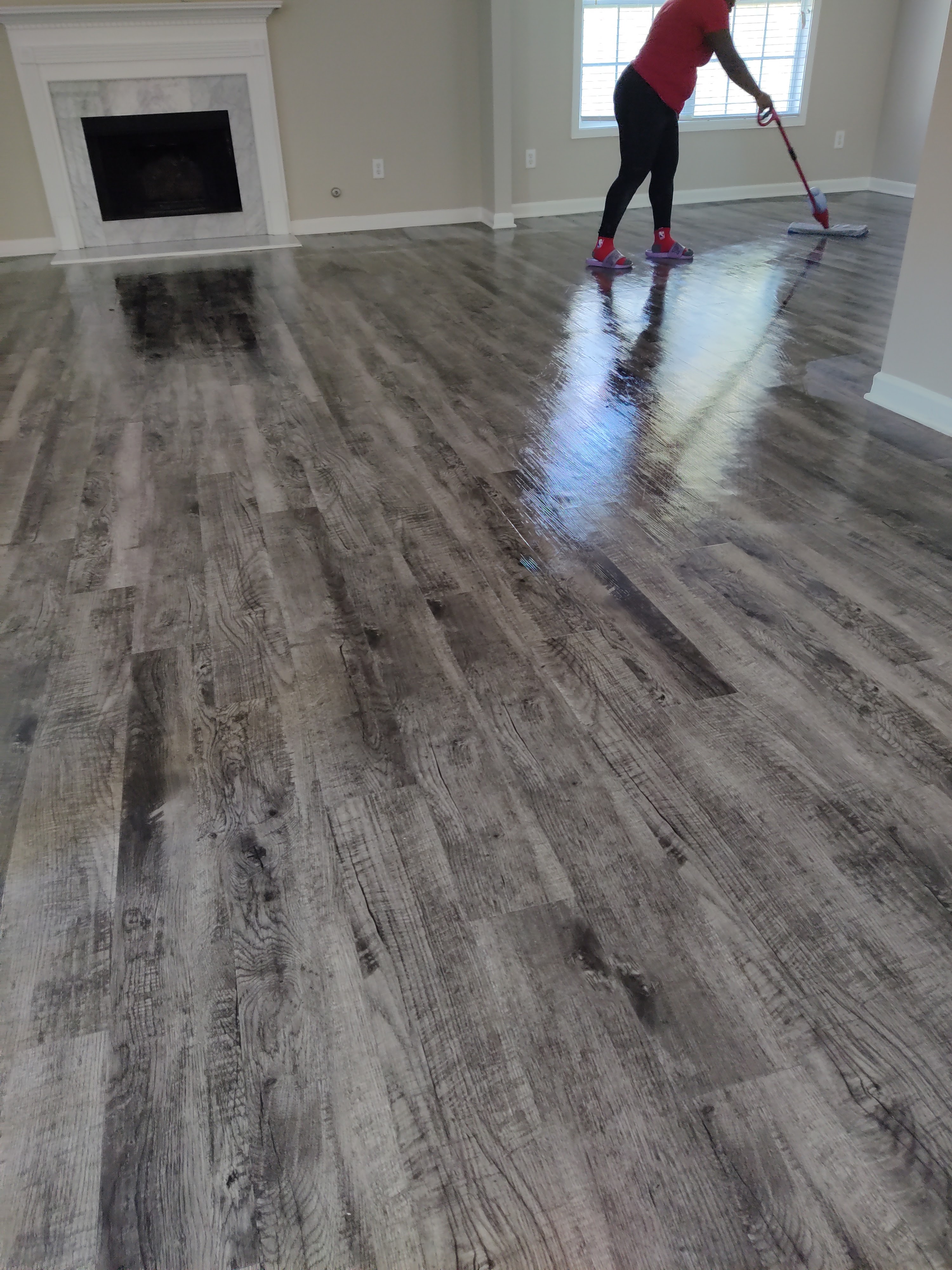 A person is mopping a shiny, grey wood-like floor in a room with a fireplace and a window. The person is wearing a red shirt, black leggings, and purple slides with red socks. The floor reflects the light from the window and appears wet.