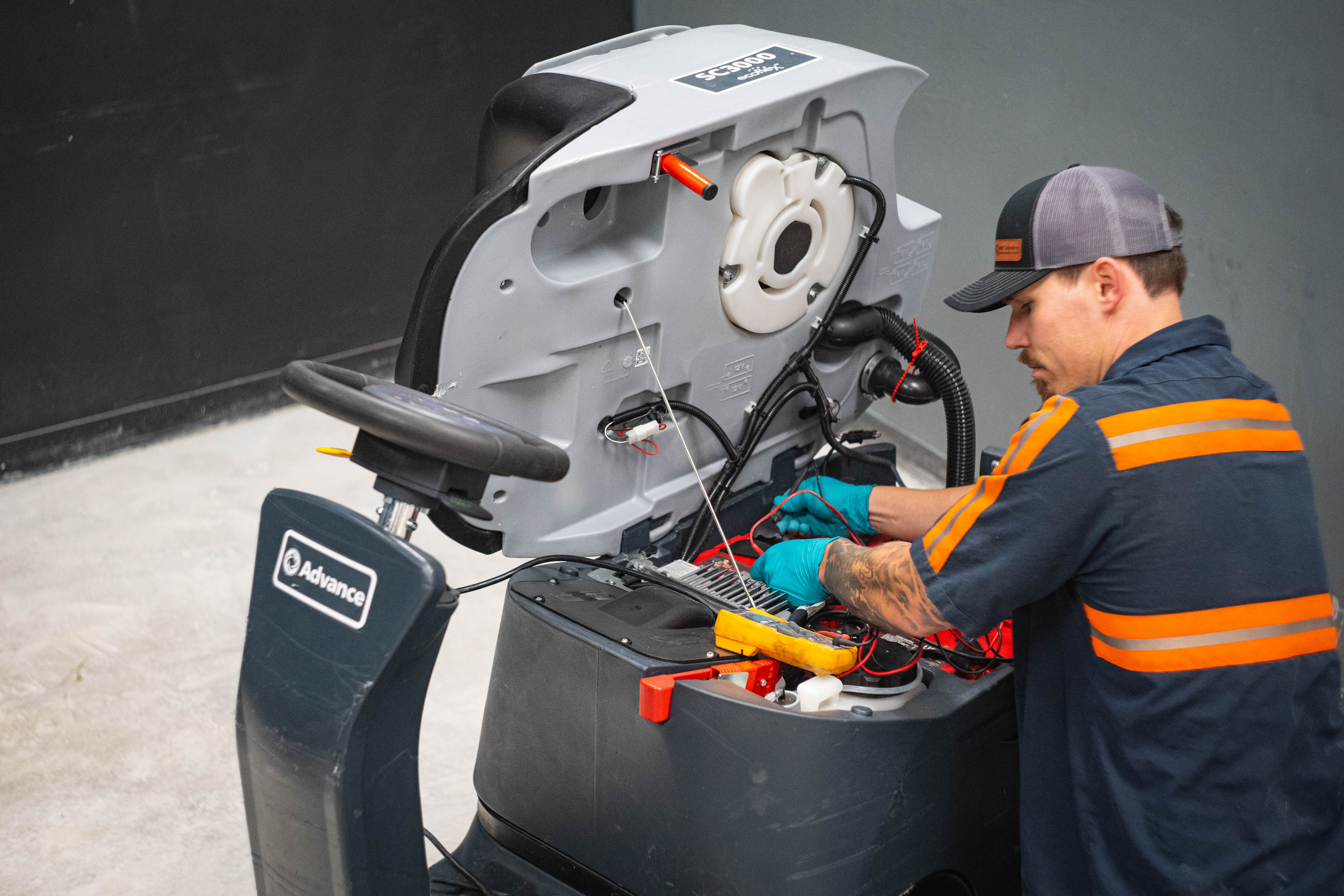 An expert service technician at WaySolutions uses a digital multimeter to test the electrical circuits of an Advance SC3000 compact rider scrubber. The machine's grey housing is open, exposing the complex wiring harness and control boards. This image highlights the technical diagnostic capabilities provided for modern computerized cleaning equipment.