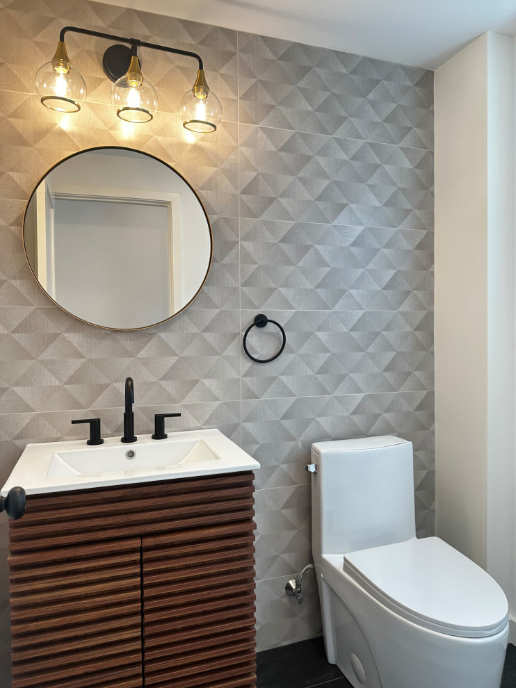 A modern bathroom features a white sink with a dark wood vanity, a round mirror, a three-bulb light fixture, and a white toilet against a textured grey tiled wall.