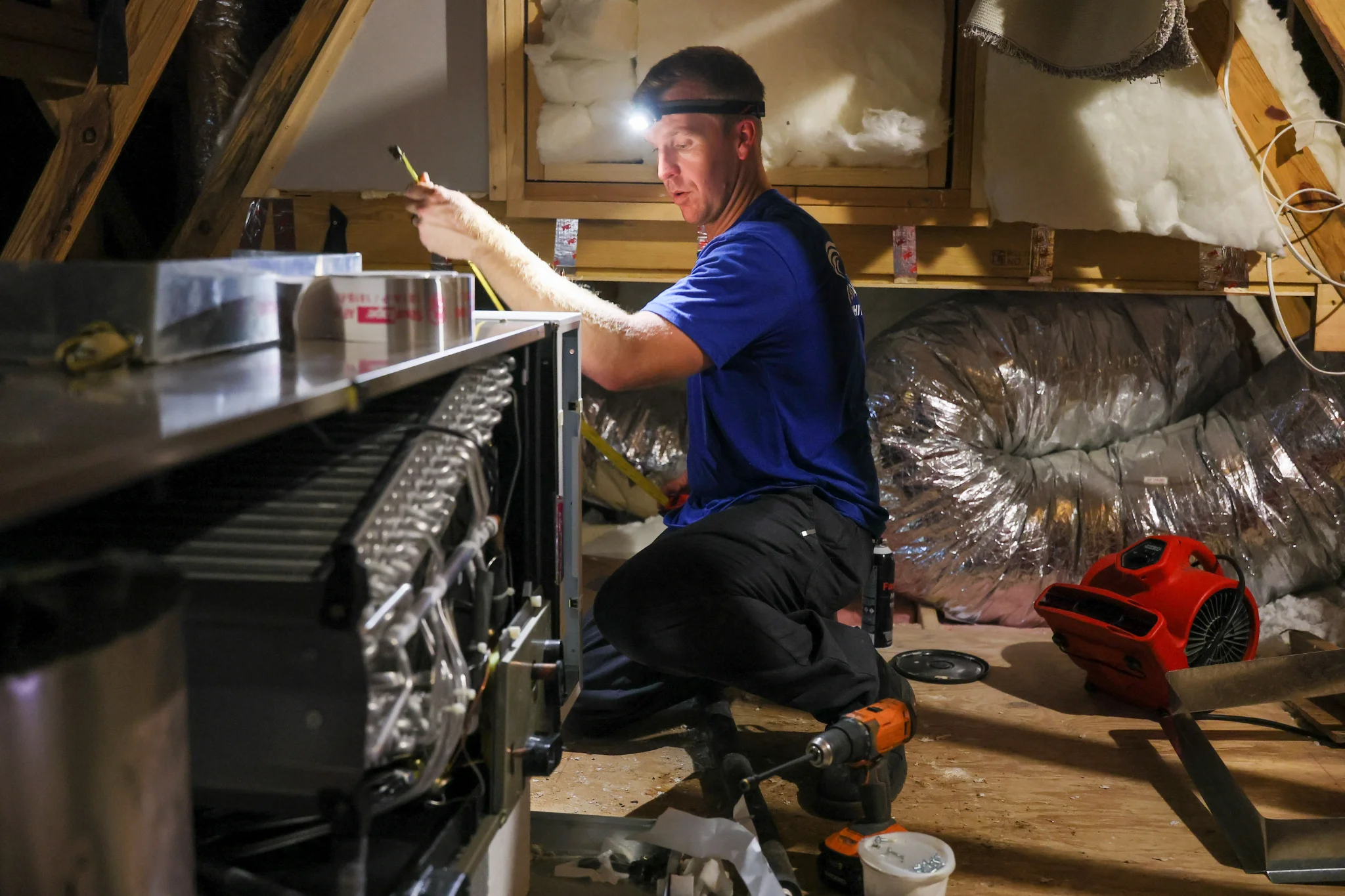 A man wearing a blue shirt and black pants, equipped with a headlamp, works on an HVAC unit in an at 