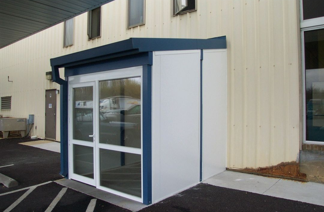 Commercial building entrance enclosure featuring glass double doors, solid side panels, and a modern blue and white frame. This vestibule-style structure provides weather protection, improves energy efficiency, and creates a clean, professional entryway for visitors and employees.