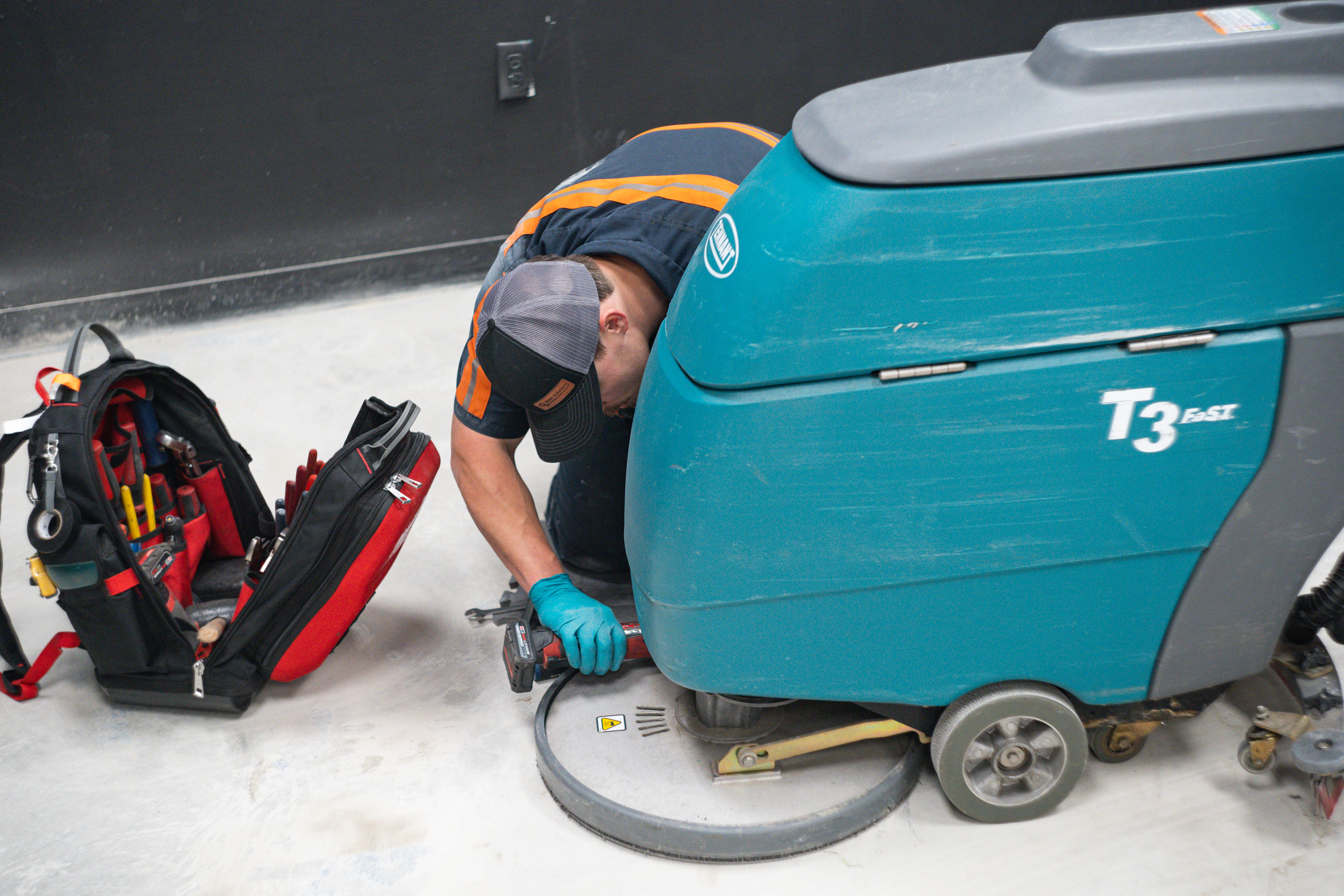 A dedicated WaySolutions technician leans over to perform deep mechanical diagnostics on the lower chassis of a burgundy Minuteman walk-behind floor scrubber. Serving industrial and retail businesses across Layton, Utah, our cross-trained mechanics ensure maximum uptime and performance for multi-brand cleaning fleets