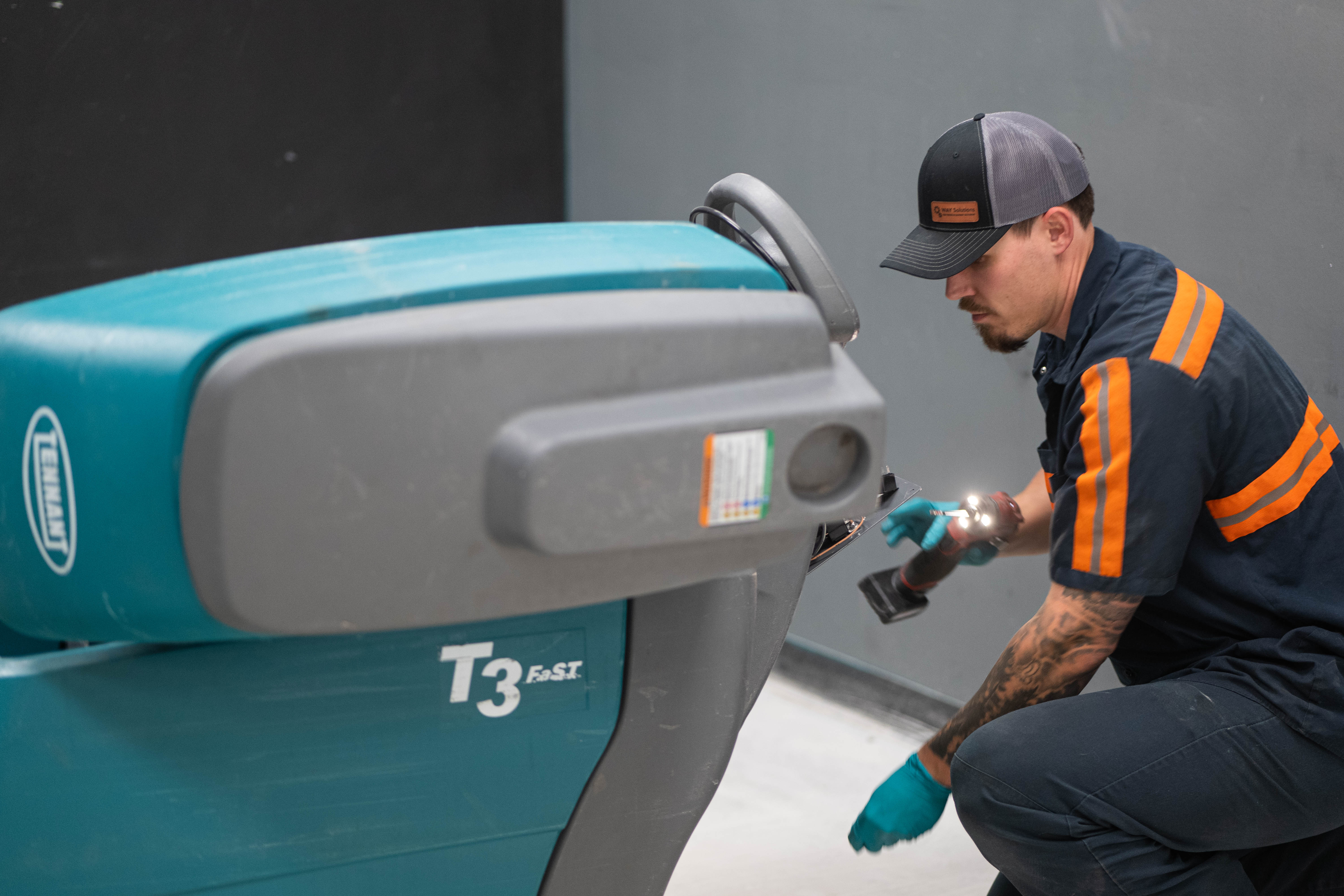 A WaySolutions service technician kneels beside a teal Tennant T3 FAST walk-behind floor scrubber, using a diagnostic flashlight and a cordless power tool to closely inspect the lower control console and steering housing. The image showcases on-site mechanical troubleshooting and expert repair capabilities for industry-standard commercial cleaning equipment.
