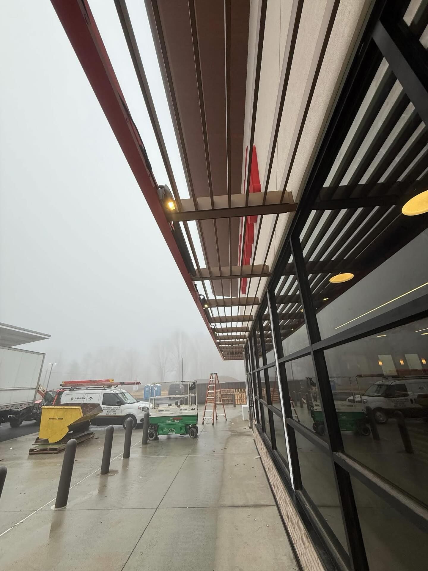 Exterior commercial building with covered walkway, construction equipment, ladder, and service vehicles visible, set in foggy weather, indicating ongoing installation or maintenance work outside retail location.