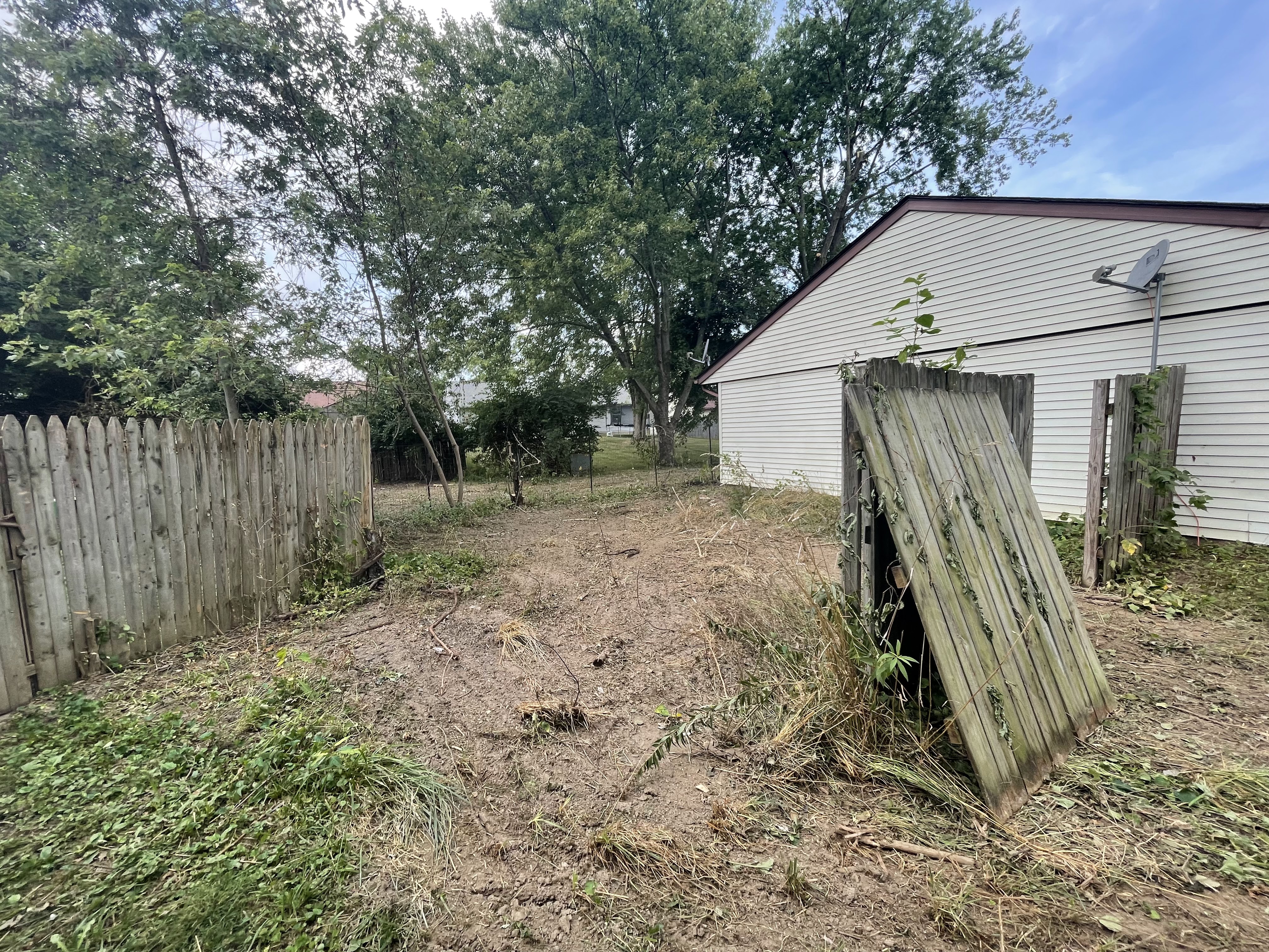 A backyard scene with a wooden fence, a white house, and trees. The ground is covered with dirt, patchy grass, and some debris. A satellite dish is mounted on the side of the house. The fence is made of vertical wooden planks and some sections appear damaged or leaning.