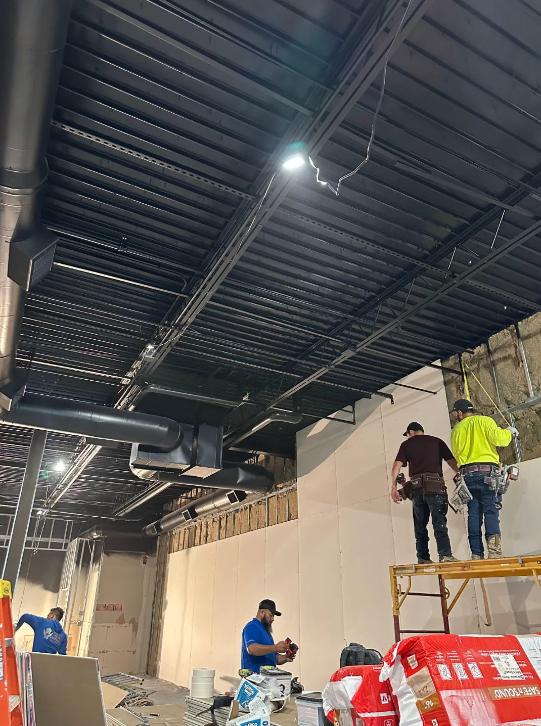 Construction crew installs drywall panels inside a commercial space using scaffolding while exposed  