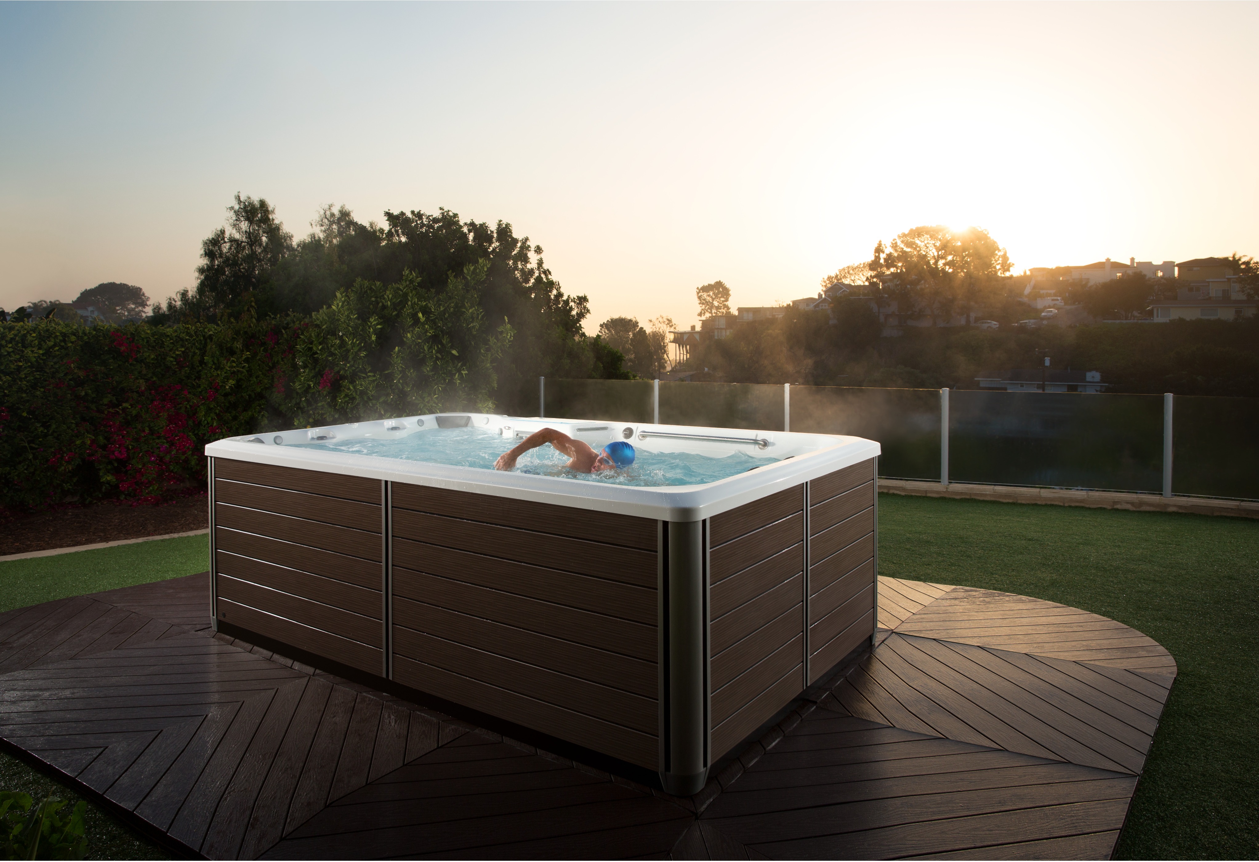 A SwimCross X200 swim spa (2016 model) in an Alpine White shell with a Dark Mocha wood-panel cabinet is shown installed on a circular hardwood deck in a residential outdoor setting. A swimmer wearing a blue cap is actively doing laps against the resistance current inside the spa. The unit is producing a visible mist, suggesting heated water. The lifestyle shot is taken at golden hour, with lush green landscaping, flowering shrubs, a glass perimeter fence, and a warm sunset sky in the background.