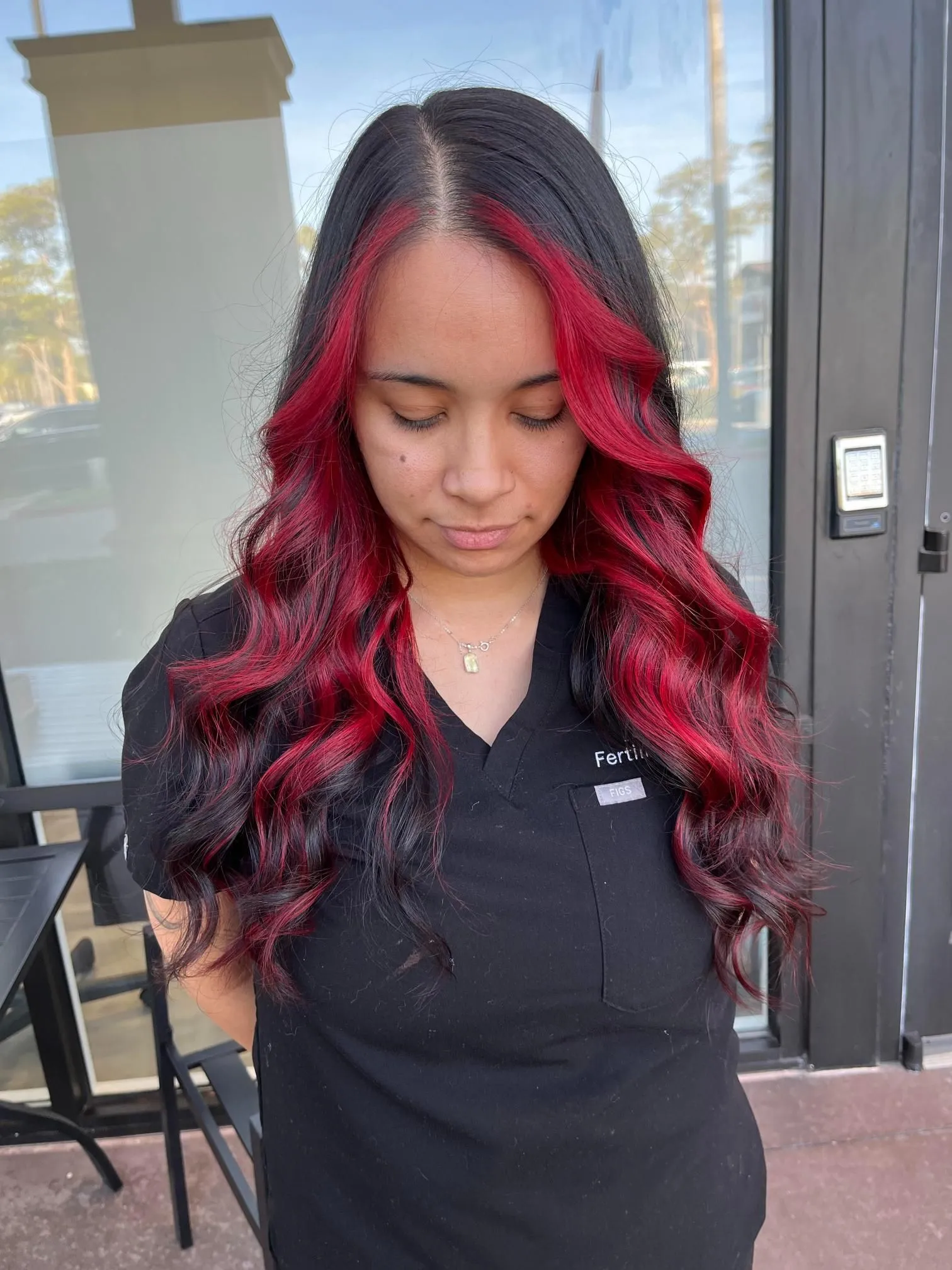Long black hair with vivid red balayage highlights styled in soft waves, creating bold contrast and dimension. The hairstyle showcases creative salon coloring techniques and professional styling photographed from the front.