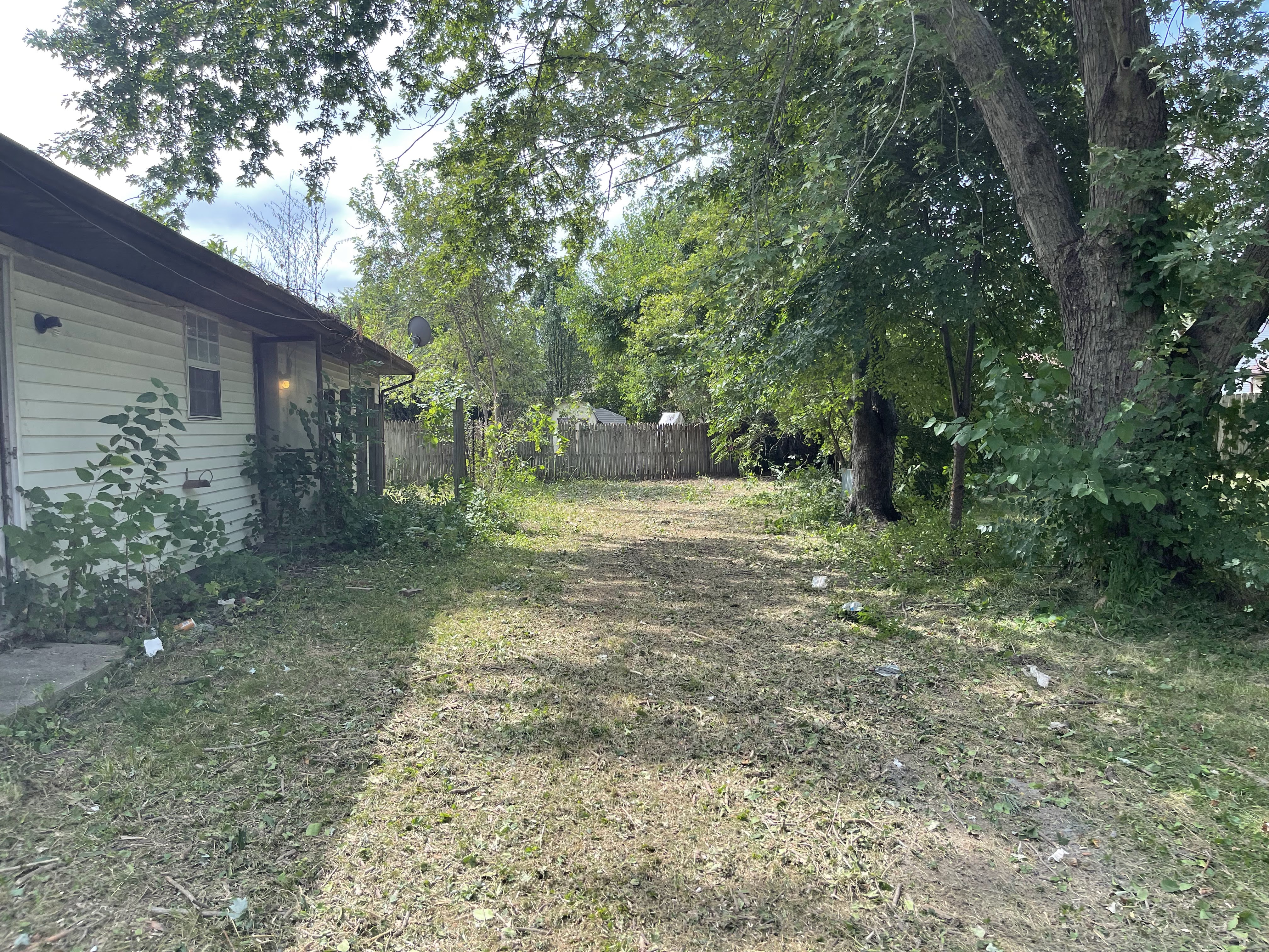 A weathered, light-colored house sits in the foreground with overgrown vegetation encroaching on its side. A wooden fence borders the yard, which is littered with grass clippings and some scattered debris. Tall trees with lush green leaves provide shade, creating a dappled sunlight effect on the ground.