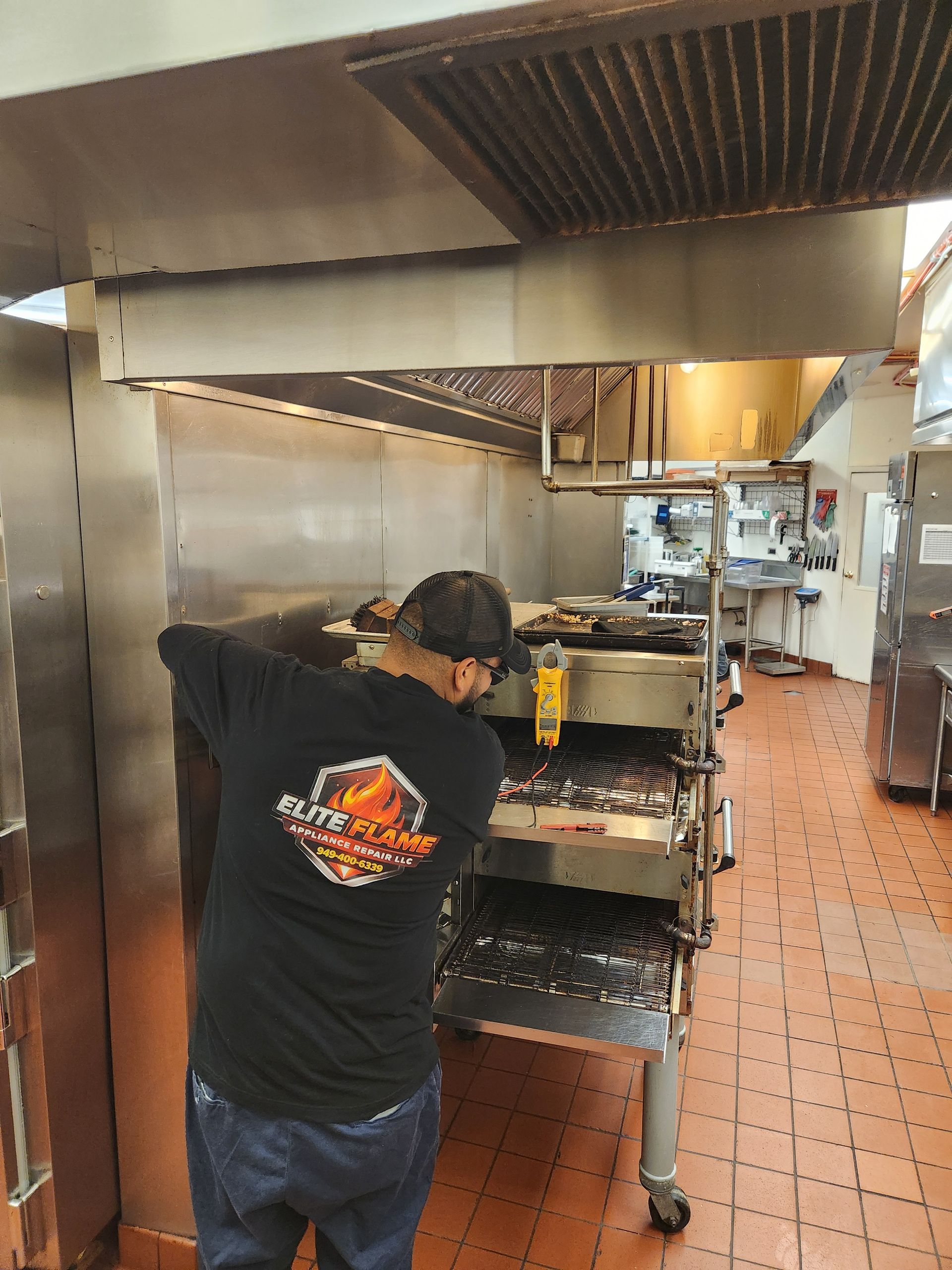 Elite Flame technician working on a commercial conveyor oven beneath a large ventilation hood, with  