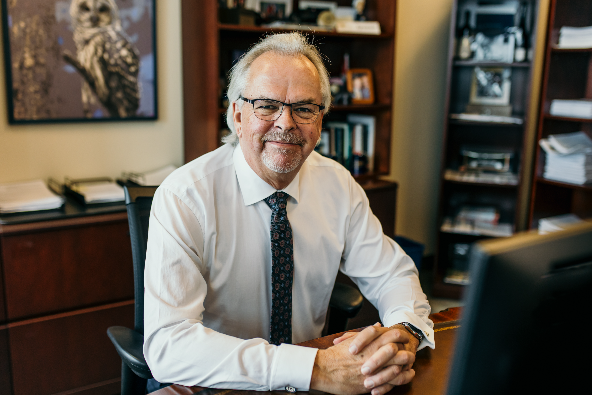 Senior Portfolio Manager & Investment Advisor Bruce MacKay seated at his desk in a professional office setting, wearing a white dress shirt and patterned tie, with bookshelves and framed photographs visible in the background.