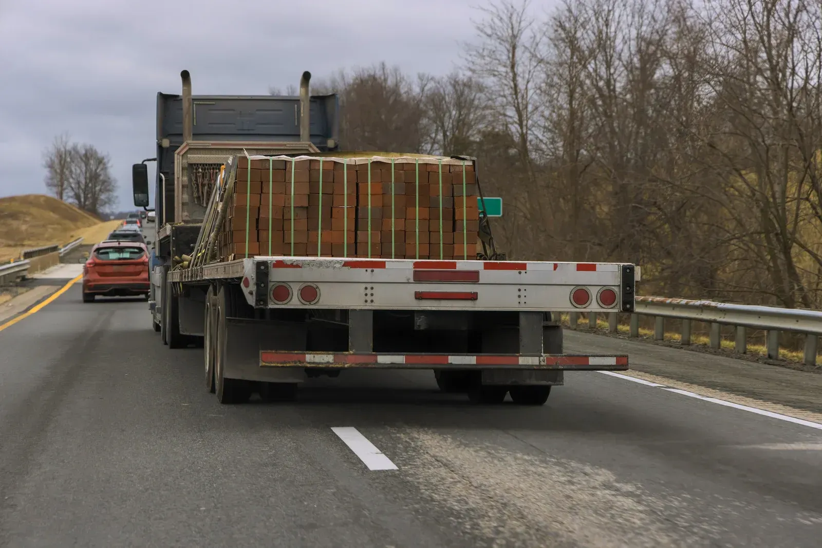 Flatbed truck transporting strapped pallets of bricks along a highway. Commercial freight delivery supporting construction projects with secure material transportation and efficient logistics coordination.