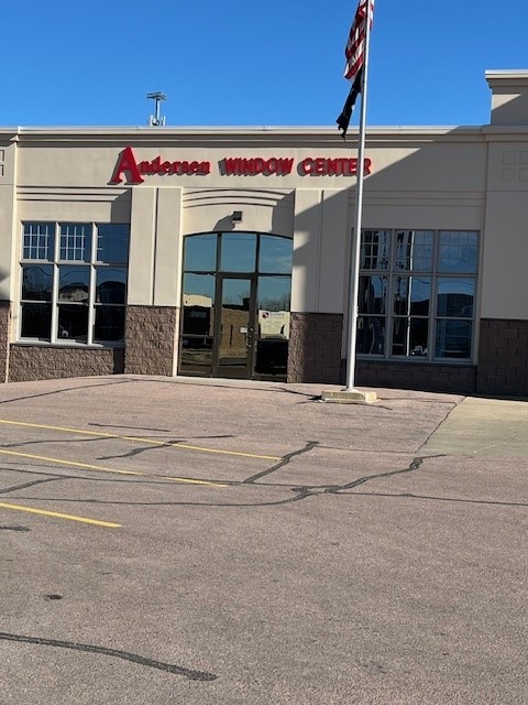 The entrance to a building labeled "Andersen Window Center" is shown, with red signage above a set of glass doors. The building has a beige and brown facade, with tall windows on either side of the entrance. A flagpole displaying the U.S. and state flags stands prominently in front of the structure. The surrounding parking lot is empty, and the photo is taken on a clear, sunny day.