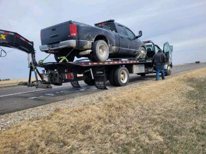 A black pickup truck is loaded onto a commercial flatbed tow truck parked along a rural highway shoulder. A person in a jacket stands near the open driver's cab door.