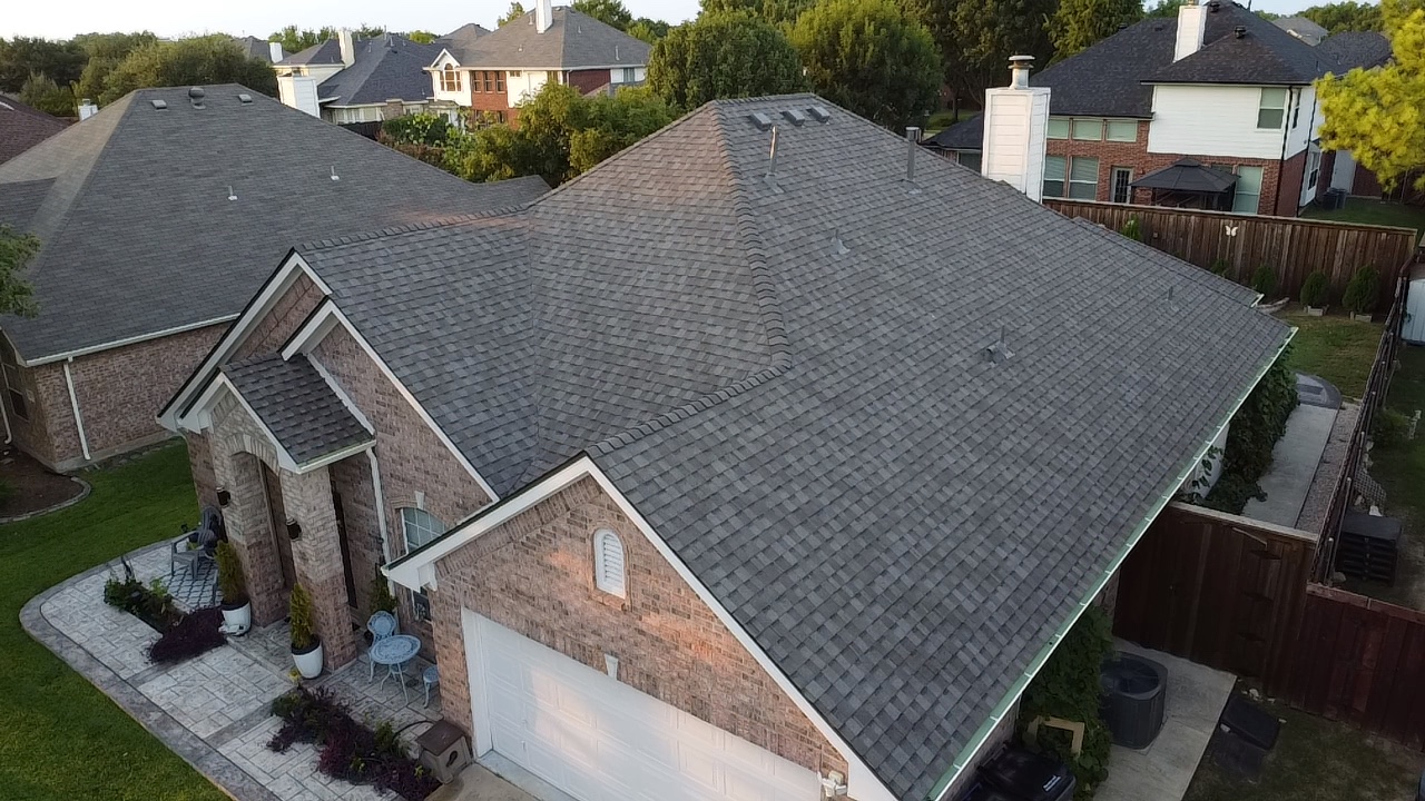 A high-angle view of a residential neighborhood with several houses and their roofs. The focus is on a brick house in the foreground with a dark gray asphalt shingle roof. The house has a white garage door and a covered entryway with brick columns. Surrounding the house are green lawns, trees, and a wooden fence.