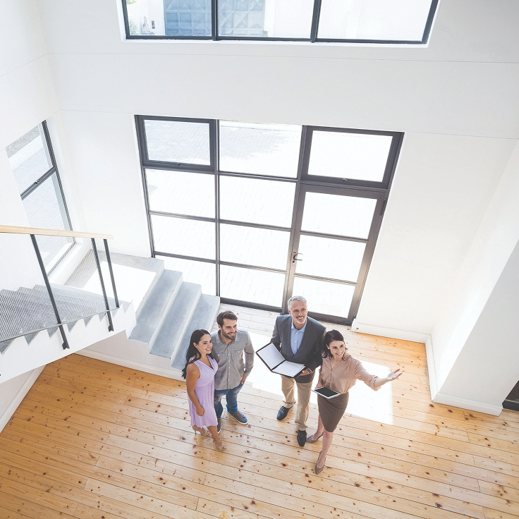 Aerial view of three people with a real estate agent inside a bright, spacious room with a wooden floor and large windows, suggesting a property tour.