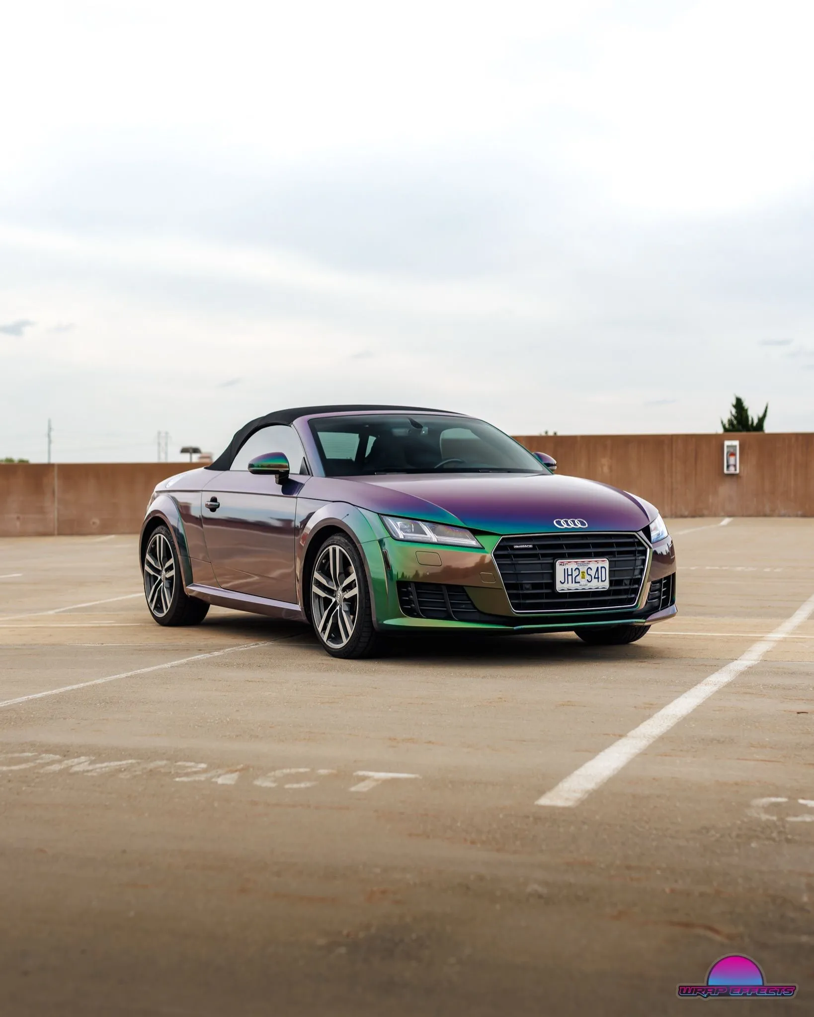 Three-quarter view of Audi TT convertible featuring premium color-changing wrap, sporty styling, black soft top, alloy wheels, and elegant automotive presentation in rooftop parking environment beautifully.