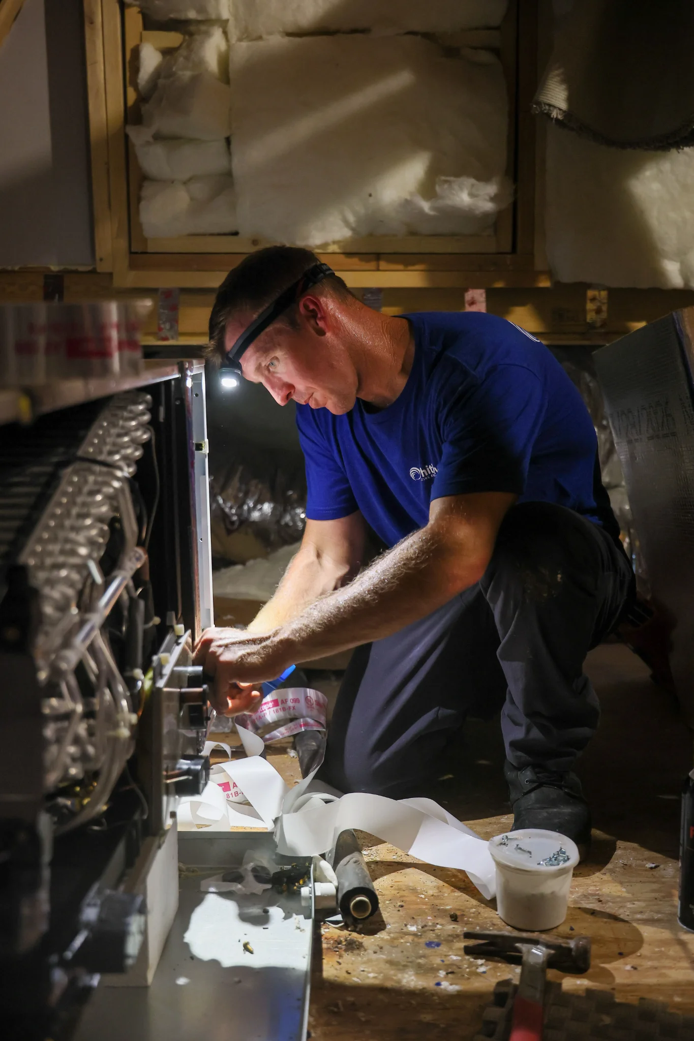 A man wearing a blue t-shirt and dark pants, equipped with a headlamp, is working on an HVAC unit in 
