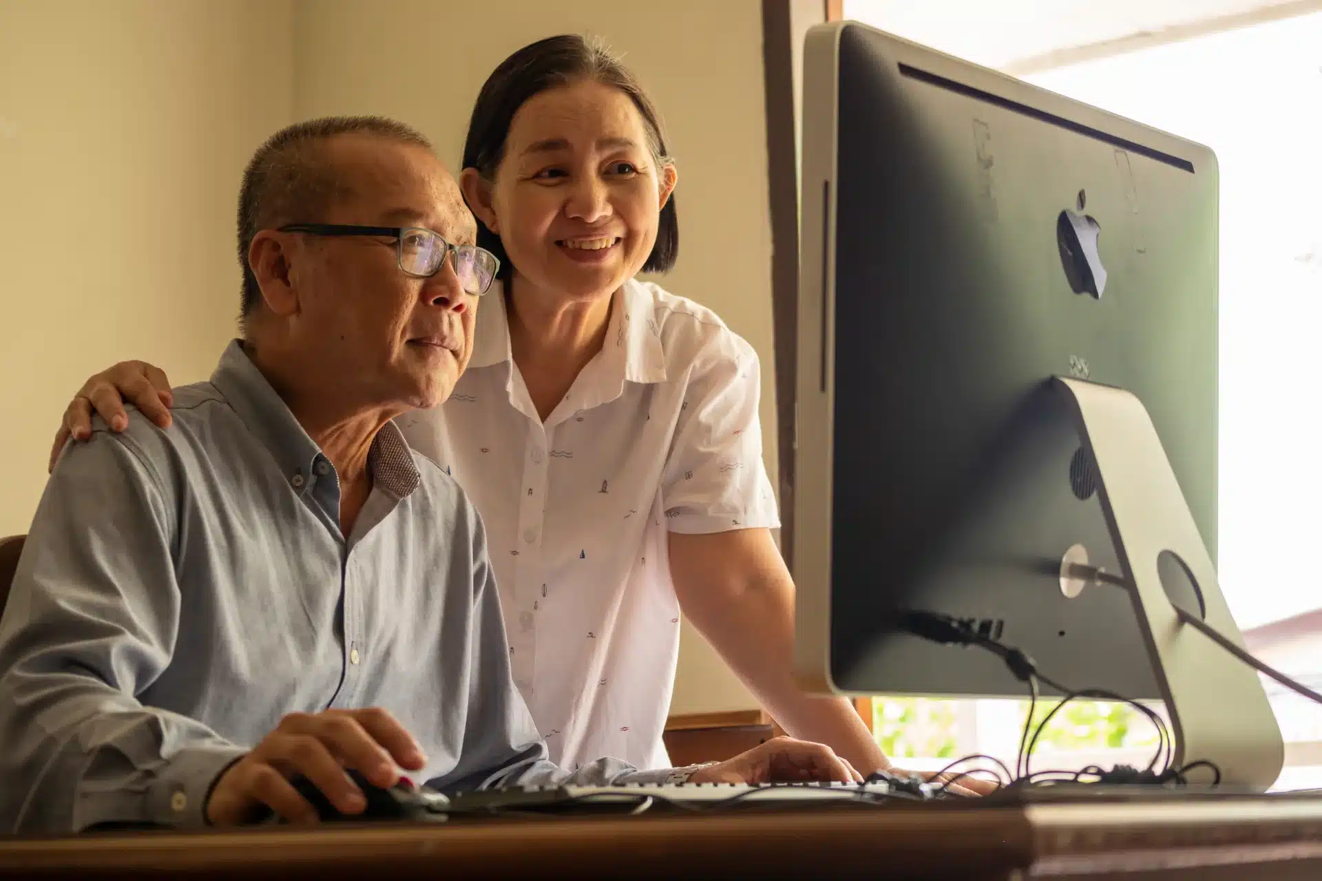 An elderly Asian couple, a man and a woman, are looking at a computer screen. The man, wearing glasses and a blue button-down shirt, is seated and using a mouse. The woman, wearing a white shirt with a pattern of small sailboats, stands behind him with her hand on his shoulder and smiles. They appear to be in a home setting, with a computer on a wooden desk.