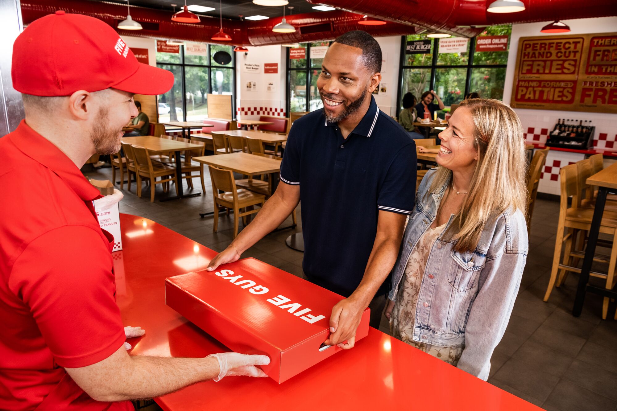 A couple collect their Five Guys catering box from a Five Guys employee at a Five Guys restaurant.