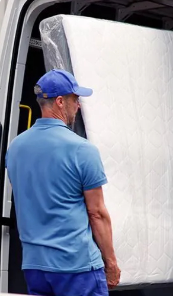 A delivery man or mover, seen from behind and wearing a blue polo shirt and a blue baseball cap, is carrying a large white mattress wrapped in plastic into the open rear doors of a white moving van or truck.