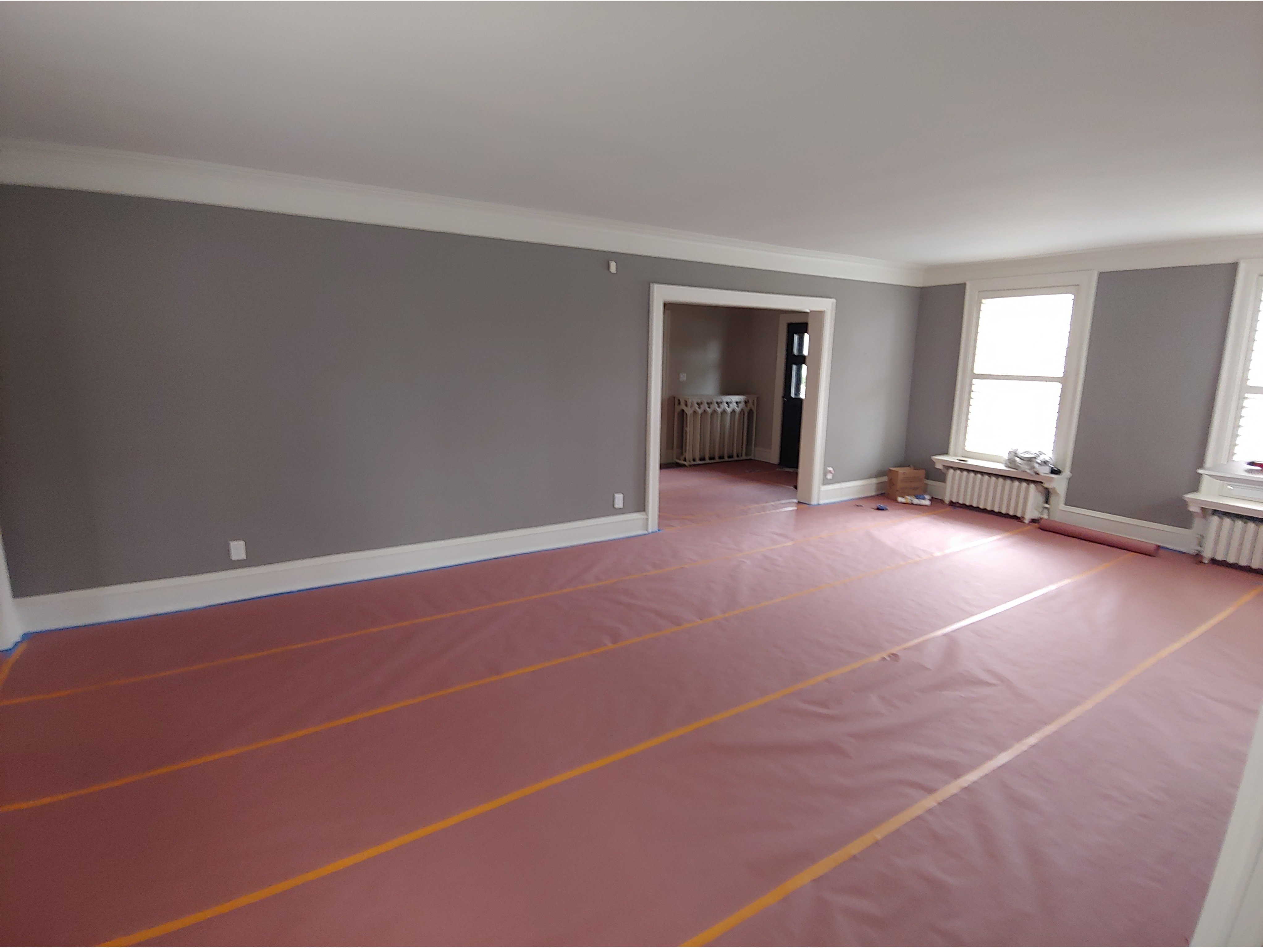 A freshly painted living room featuring neutral gray walls with white trim and crown molding for a clean, modern look. The floors are protected with red covering during the painting process, highlighting professional preparation and attention to detail. Large windows with white trim provide natural light, making the space bright and inviting. Ideal for showcasing interior painting services, wall prep, and renovation projects.