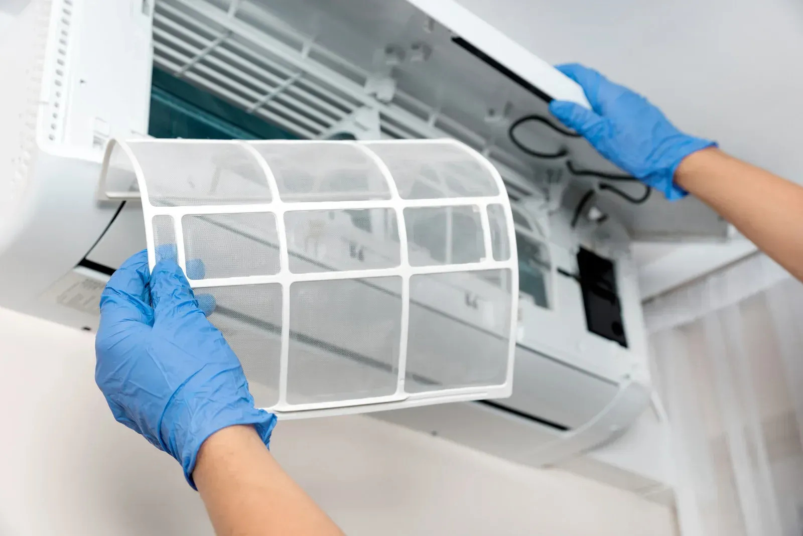 A person wearing blue gloves is removing or replacing a white mesh air filter from the front panel of a white, wall-mounted mini-split air conditioner unit.