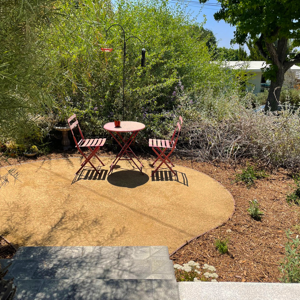 Sunny garden patio with two red folding chairs and a round table, surrounded by lush greenery and casting crisp shadows in a welcoming space.