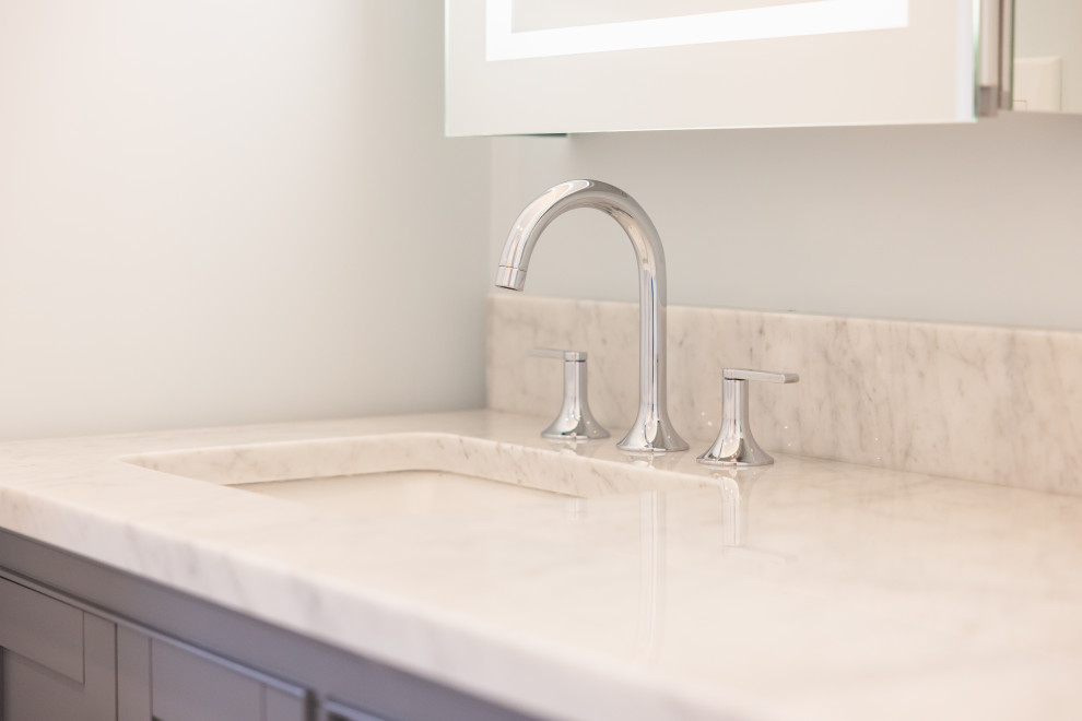 A close-up shot of a modern bathroom vanity featuring a sleek chrome faucet with two matching handles, set against a white marble countertop and backsplash. A lit mirror is visible at the top, reflecting light.