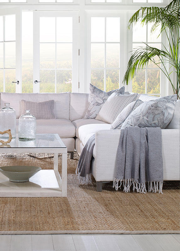 A coastal themed living room with large windows, a sectional couch, and a natural woven grass rug.