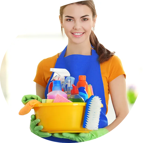 A smiling woman in an orange t-shirt and blue apron holds a yellow bucket filled with cleaning supplies, including various spray bottles, sponges, and a scrub brush. She is wearing green gloves.