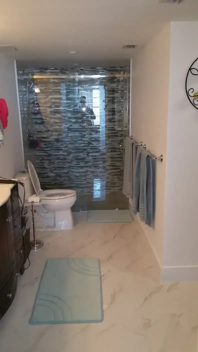 A bathroom features a white toilet with the lid and seat open, situated near a glass-enclosed shower. Towels hang on a rack, and blue bath mats are on the floor.