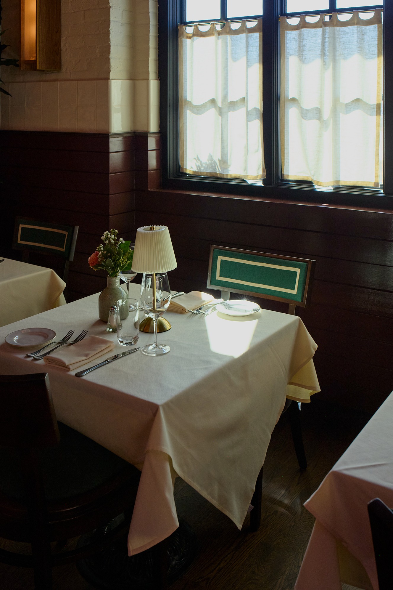 A cozy restaurant corner featuring a white-clothed table and a leather booth. Sunlight streams across the scene, highlighting a large green industrial pipe and a soft yellow cone-shaped pendant light.