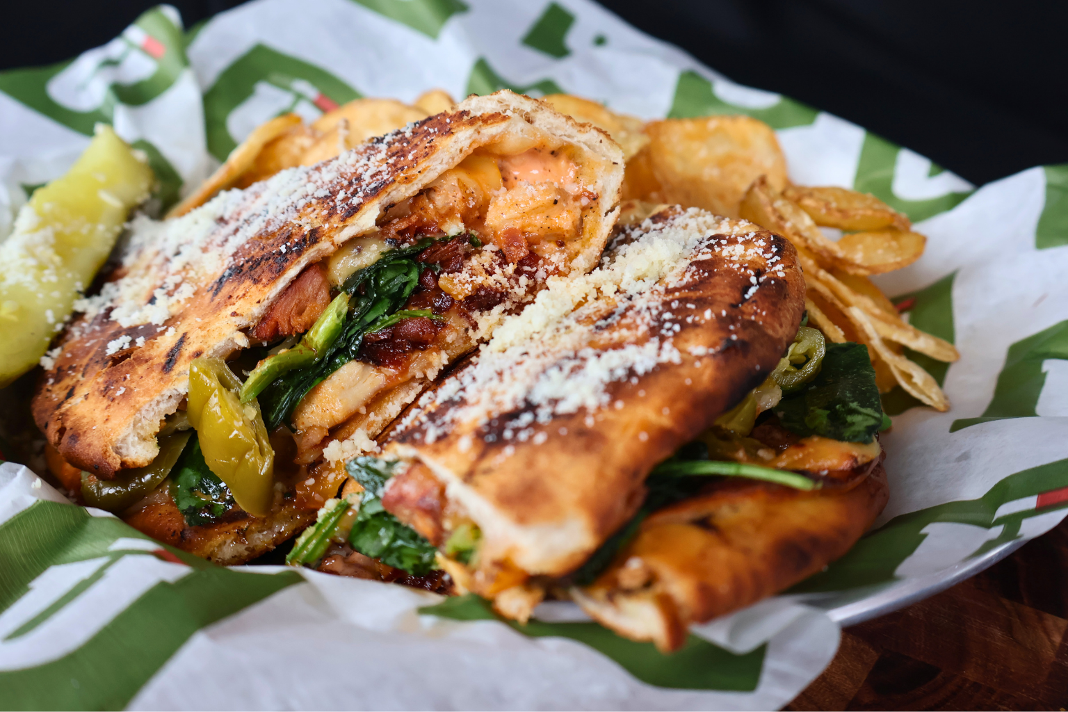 Close-up of a toasted flatbread sandwich showing a cross-section of chicken, gouda, and spinach. It is dusted with cheese and paired with golden chips on green-and-white deli paper.