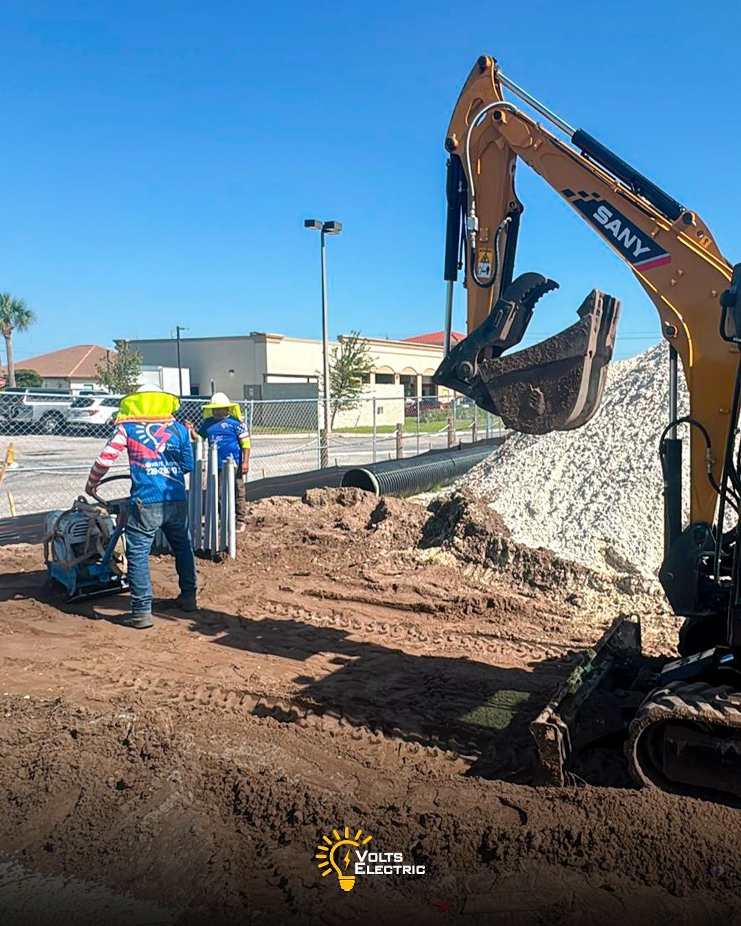 Construction workers installing underground electrical conduits at a commercial site while compacting soil near heavy machinery and a large excavator, preparing the ground for electrical infrastructure installation.
