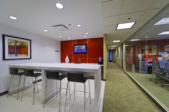 A stylish office lounge area featuring a sleek white high-top table with four modern black barstools. The lounge is accented by a bold red wall with a mounted TV displaying news and a decorative white vase adding a minimalist touch. Adjacent to the lounge, a glass wall offers a view into a conference room, and a carpeted hallway leads to additional office spaces. The neutral tones of the walls and flooring complement the vibrant red feature wall.