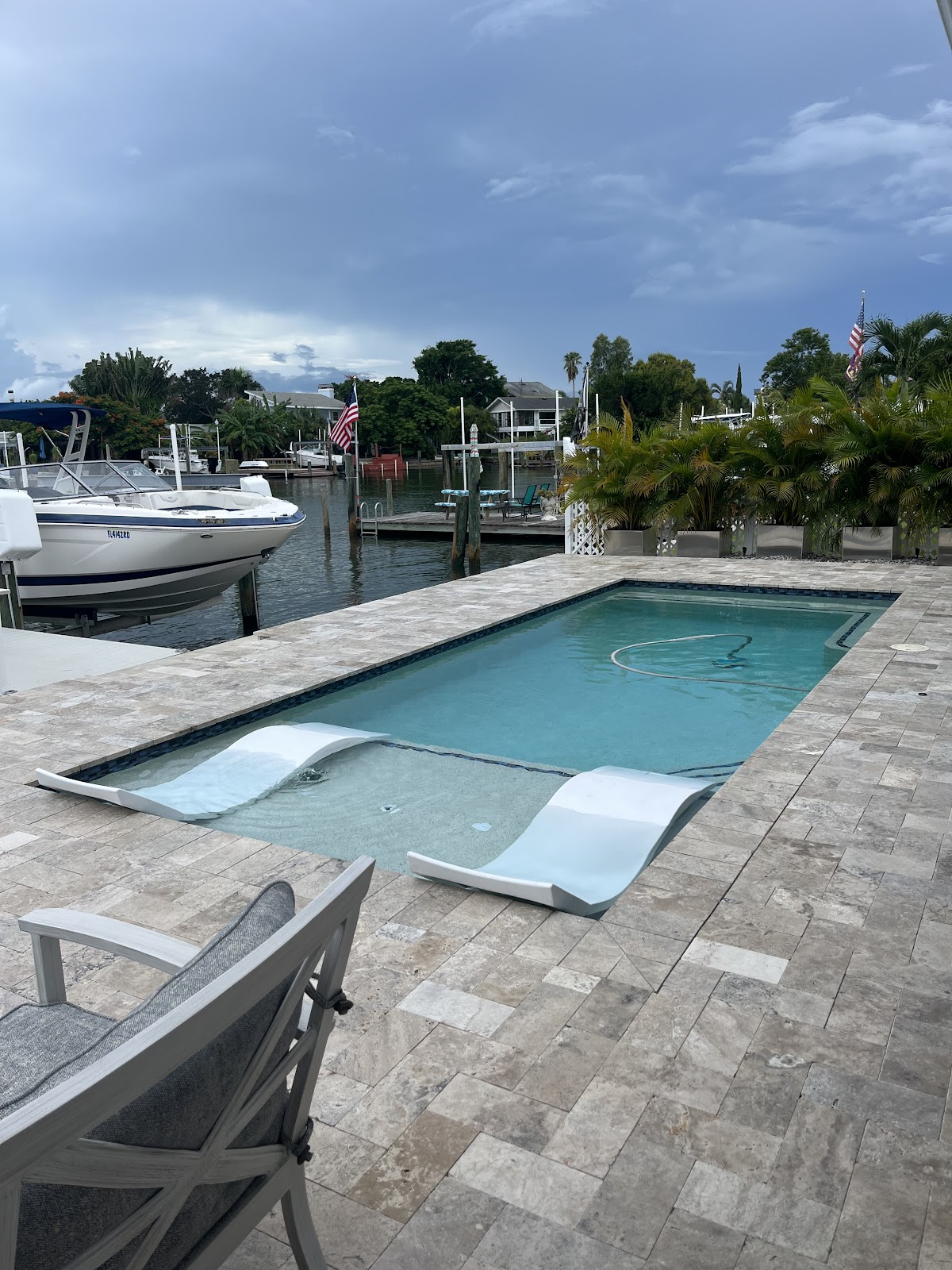 Modern waterfront backyard pool with a shallow sun shelf, in-pool lounge chairs, stone paver decking, and dock access.