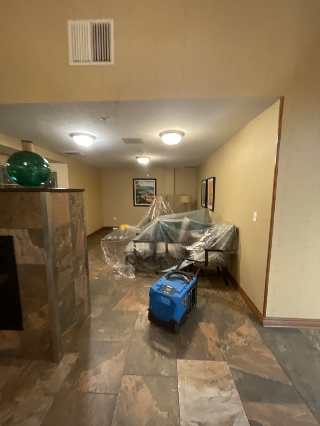 A living room with furniture covered in plastic sheeting and a blue dehumidifier in the foreground. The room has slate tile flooring, a fireplace on the left, and artwork on the walls. Several lights are visible on the ceiling.